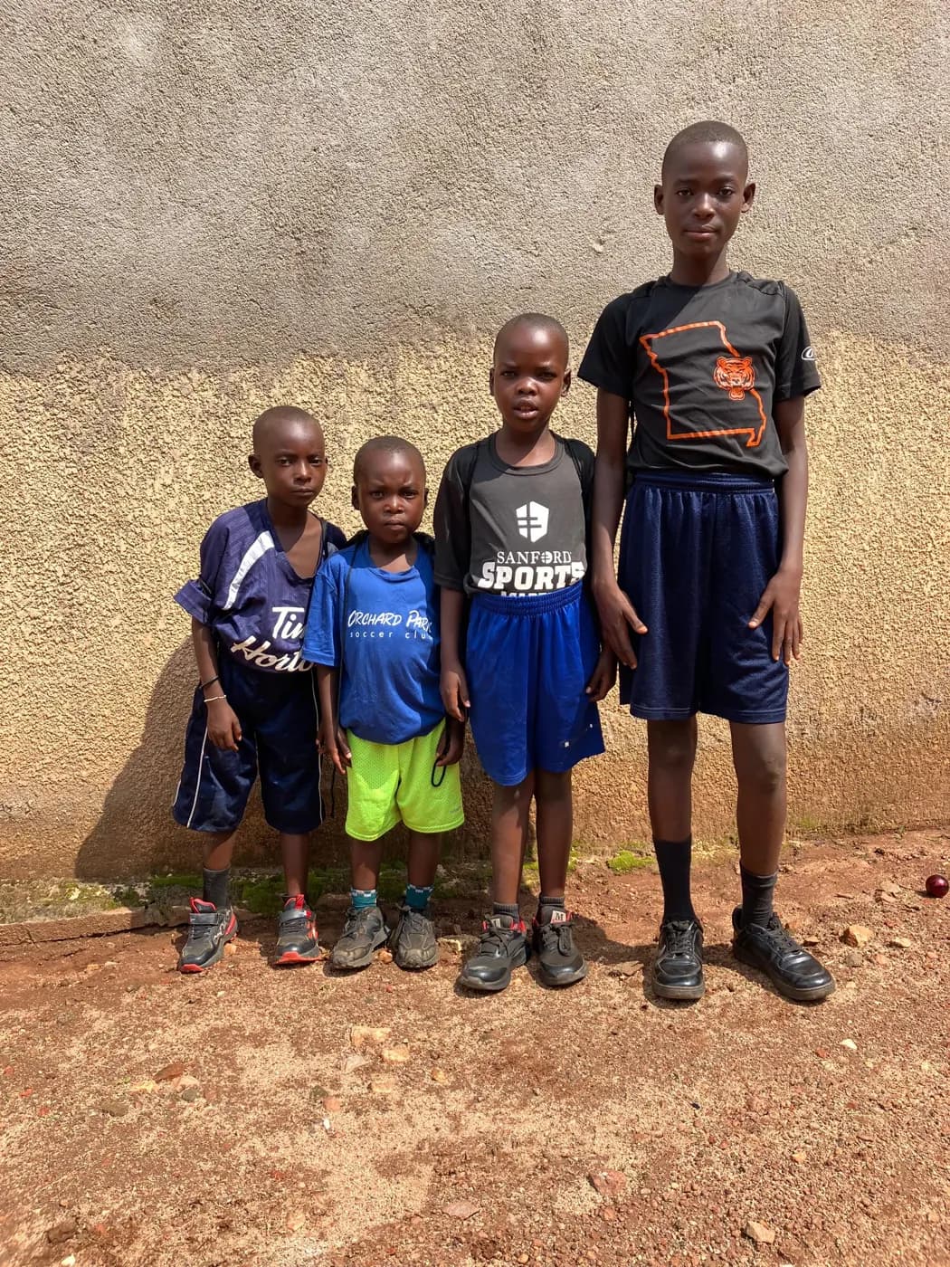 Four children standing together outside a home, wearing shoes and everyday clothes