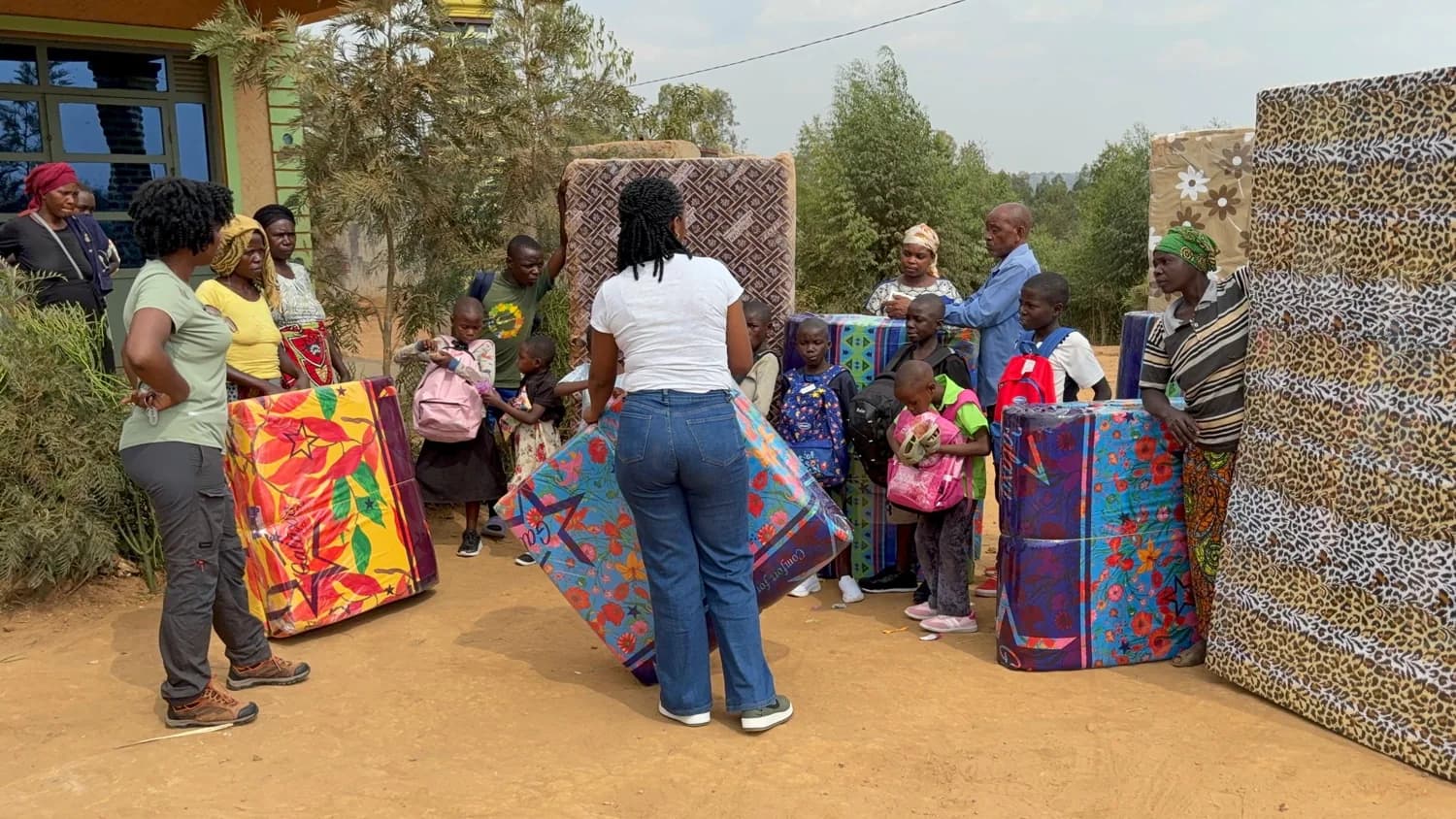 Children and adults gathered outdoors with backpacks and upright mattresses